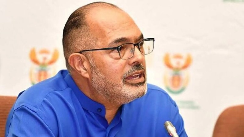 A man with glasses speaking during a press conference, wearing a blue shirt, with a backdrop featuring the emblem of South Africa.